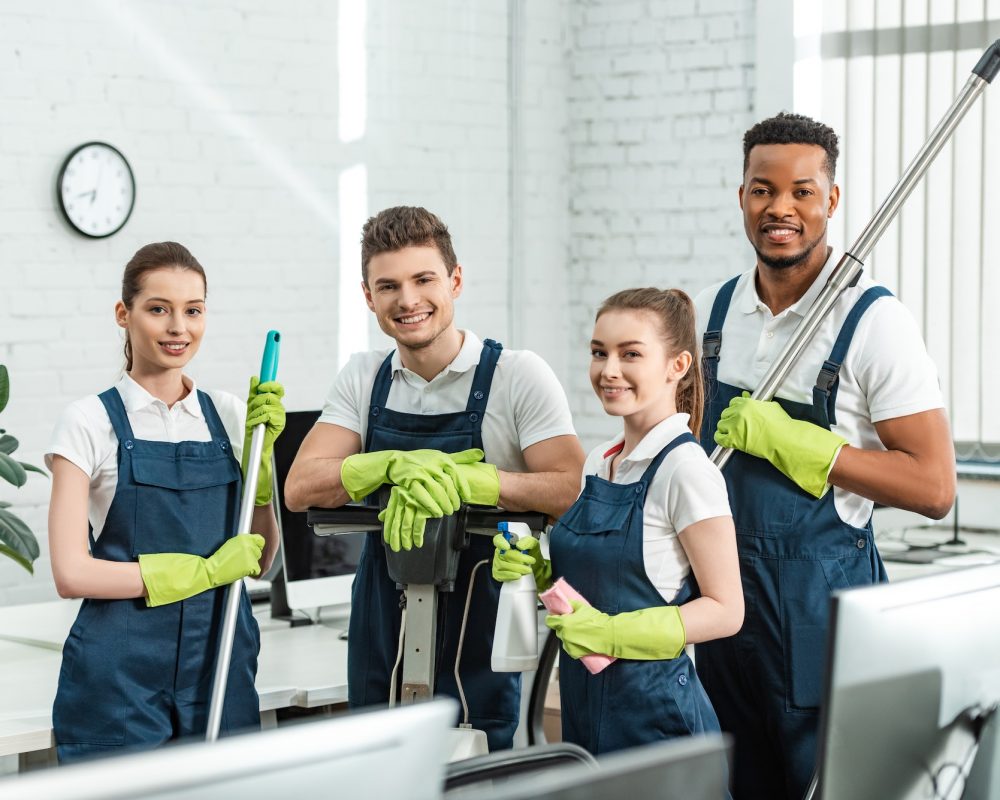 happy multicultural cleaners looking at camera while standing with cleaning supplies in office